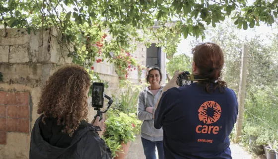 Back of two women wearing dar blue jackets with CARE's orange logo filming a woman wearing gray jumper, smiling to the camera