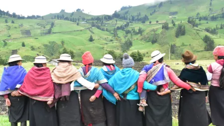Women standing outdoors, facing away from the camera, standing in a line, arms linked or around each other. 