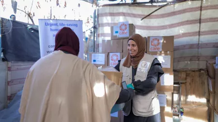 Woman wearing brown headscarf handing in box to a person. Boxes with CARE and EU logos are piled up in the background.