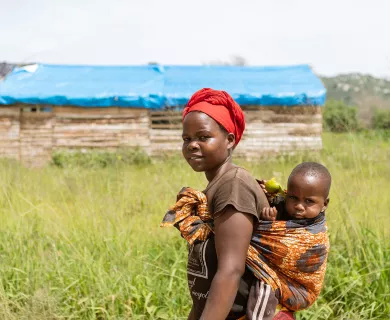 A woman stands in a field, with a young child rest against her back. He is swaddled in a patterned cloth that is tied around the woman's chest.