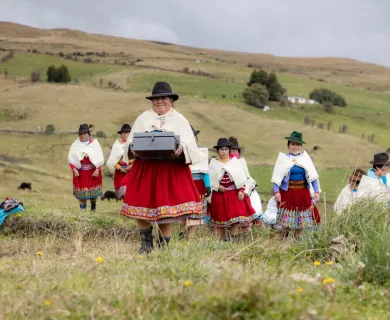 Women in typical educadorian outfits walking in a field. Woman in the forefront carrying a closed box